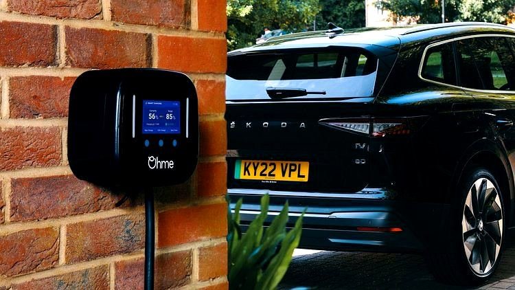 Ohme Home Pro EV charger mounted on brick wall showing colour LCD screen charging status beside black Skoda Enyaq