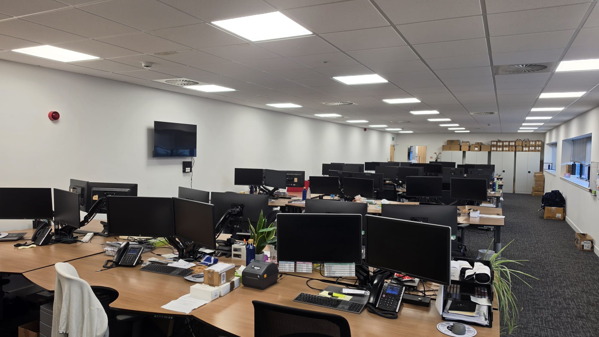 Open plan office with rows of desks and monitors lit by recessed LED panels across a suspended ceiling